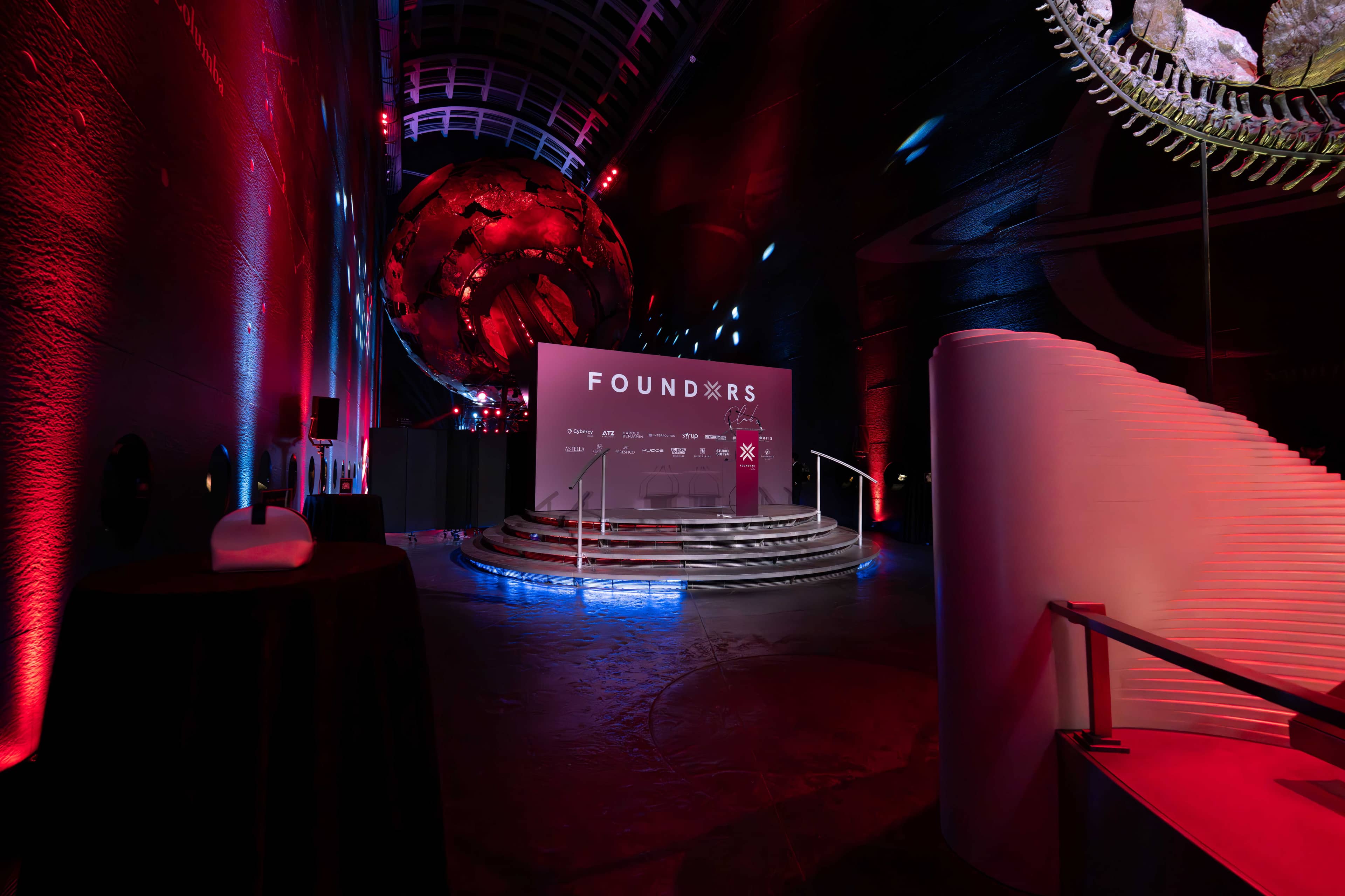 The printed FOUNDXRS stage backdrop in Earth Hall at the Natural History Museum, red uplighting on the walls and blue light along the curved balconies, printed and lit by Kudos AV